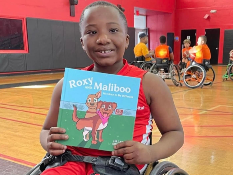 Child in a wheelchair holding the 'Roxy and Maliboo' book at a basketball court with other children and adults in the background.