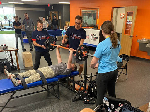 GLASA athlete performing a bench press with 25lbs on each side while their thighs and ankles are strapped to the bench with volunteers spotting the lift.