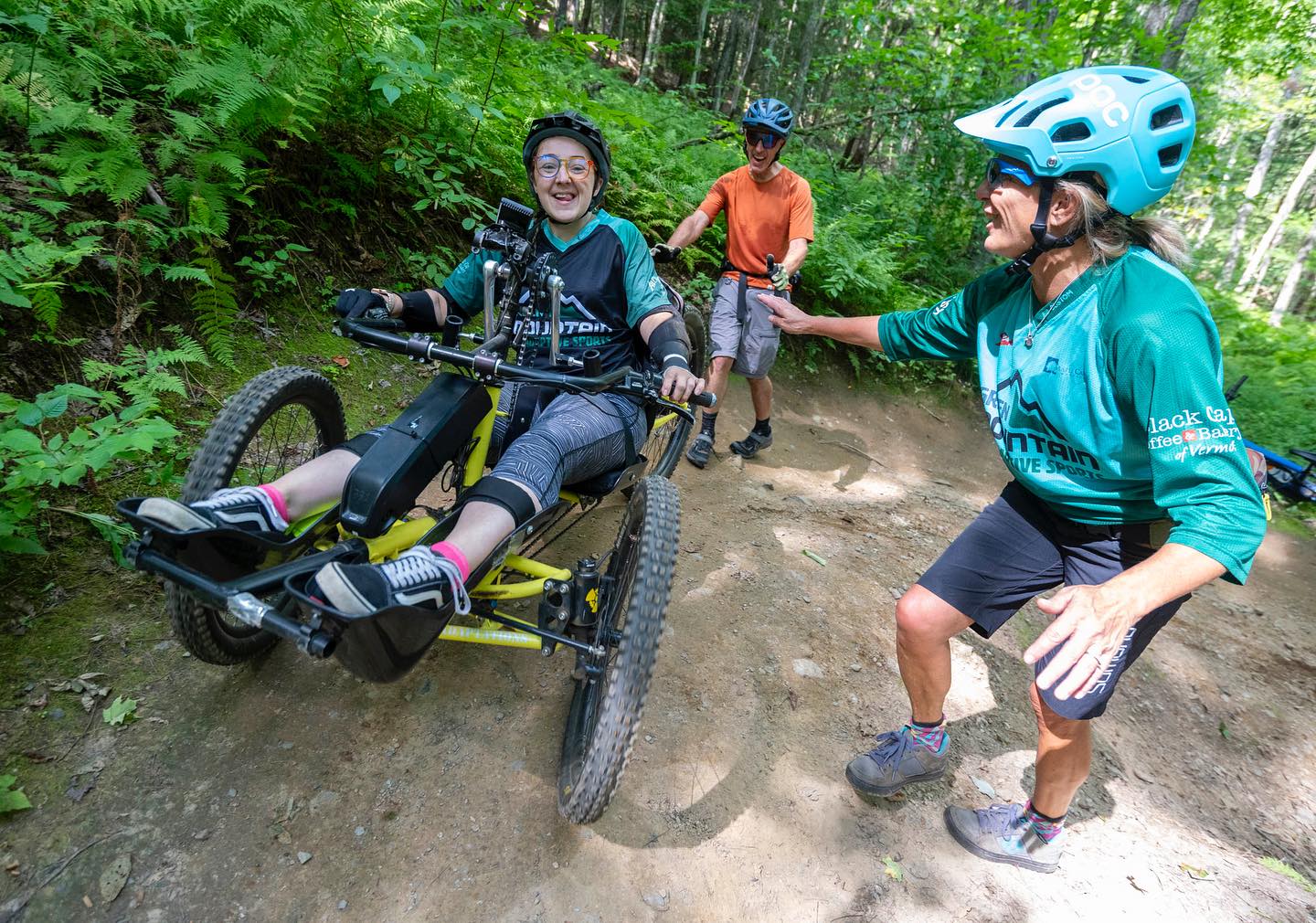 GMAS participant riding an adaptive mountain bike with assistance from people on a forest trail.
