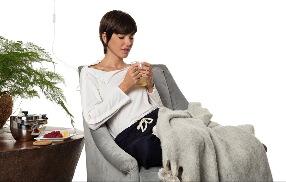 Woman sitting in a chair wearing the MAI We Care Women's Adaptive Long Sleeve T-Shirt with the collar unbuttoned to allow access to her IV, while holding a mug with a plant and table in the background