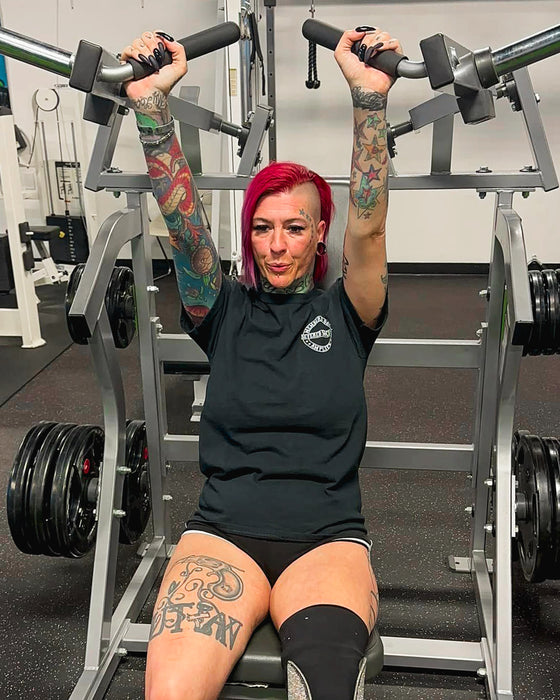 SuZanne Jacobsen performing a machine shoulder press while wearing the Severed Society® Members Only T-Shirt.