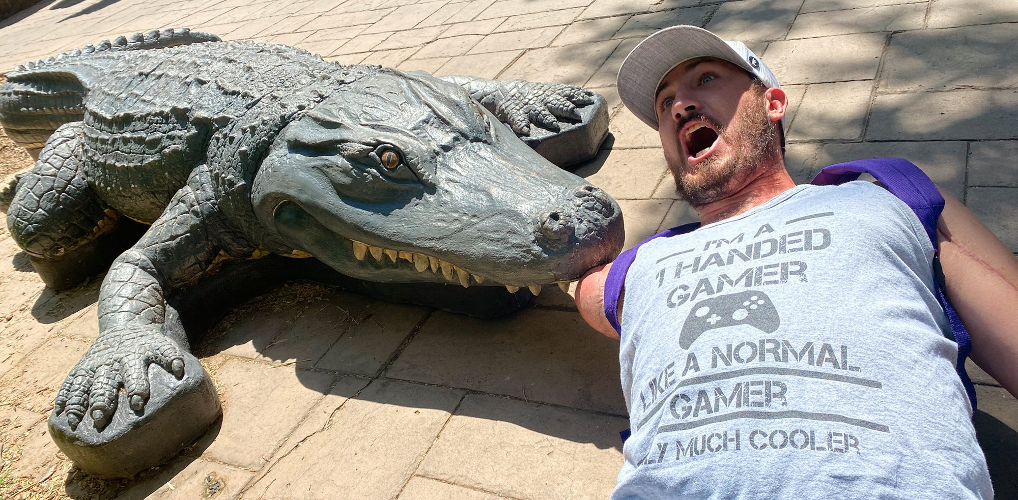 Aaron Soetaert laying on his back with his right shoulder disarticulation amputated arm in the mouth of an alligator statue pretending that the alligator bit his arm off.