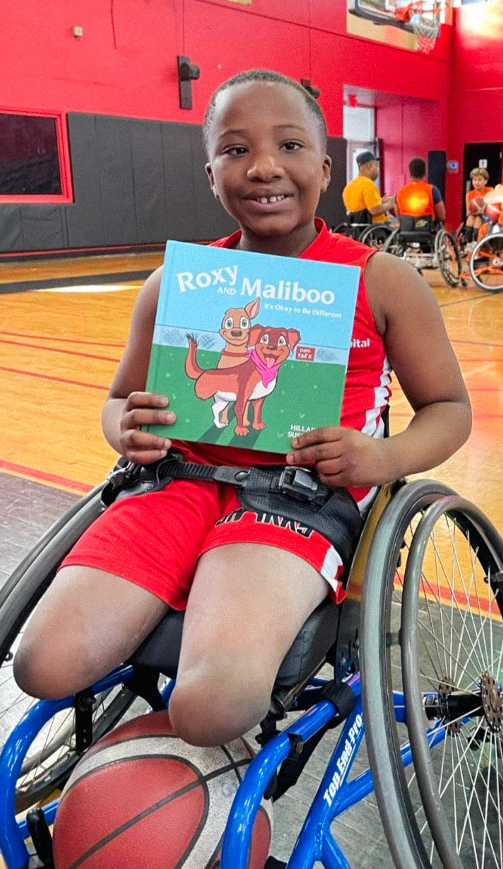 Child in a wheelchair holding the 'Roxy and Maliboo' book at a basketball court with other children and adults in the background.