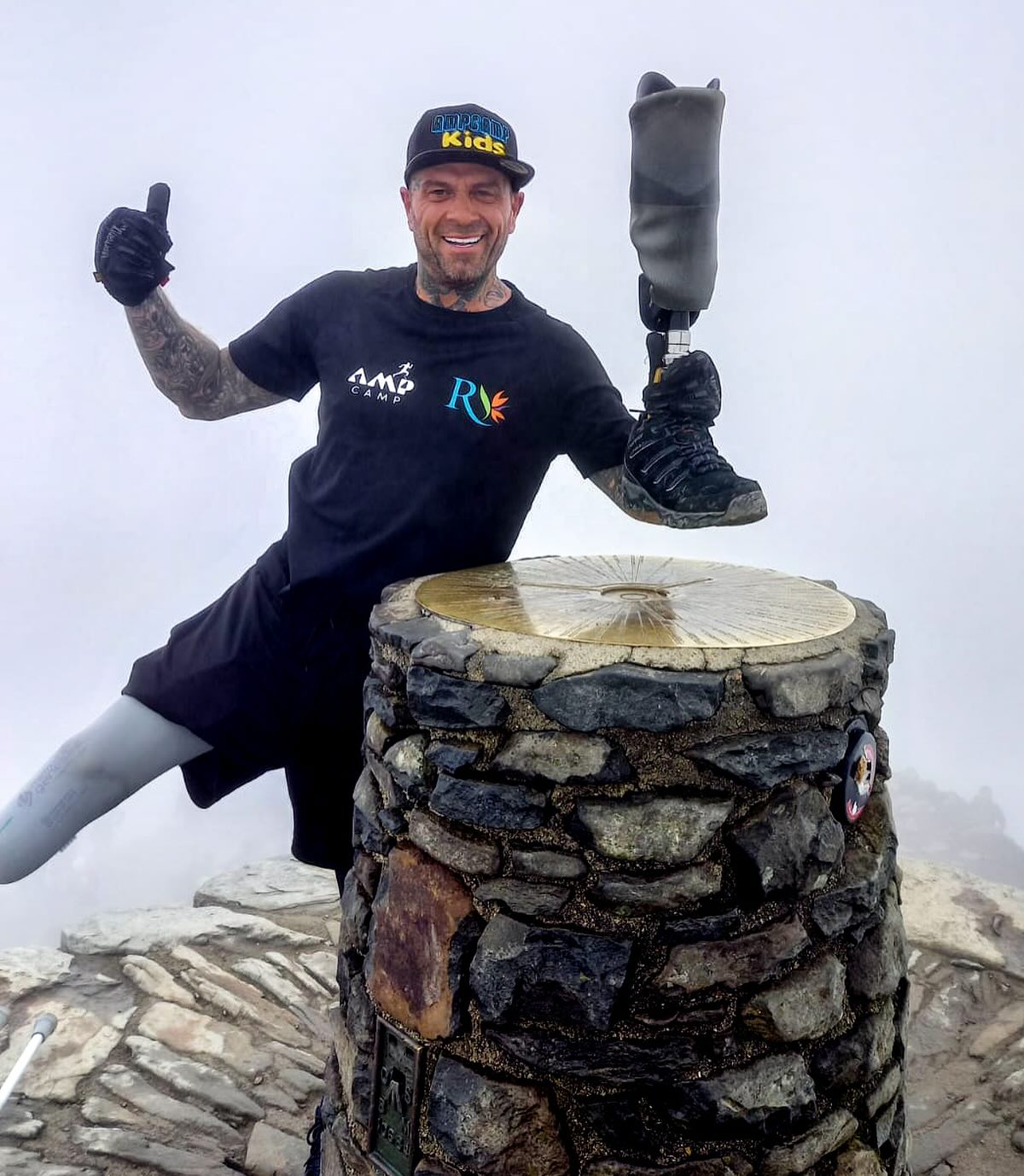 Team Amplife® Ambassador Ben Lovell holding his prosthetic leg while giving a thumbs-up on a mountain summit