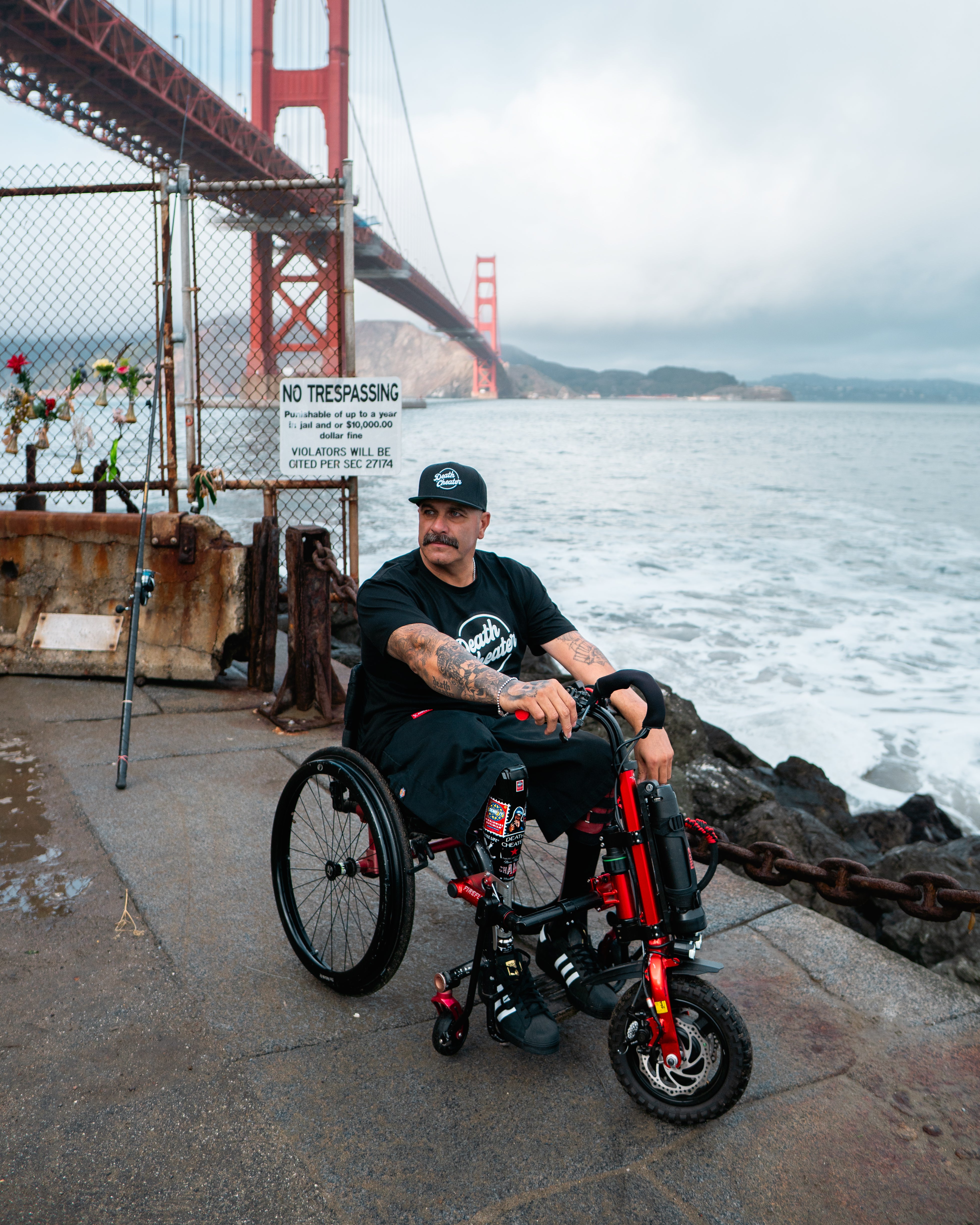 Amplife Founder Abdul Nevarez riding his Rio Mobility Firefly 2.5 at the edge of Fort Point under the Golden Gate Bridge while wearing the Death Cheater® Halo T-Shirt and Flat Bill Snapback.