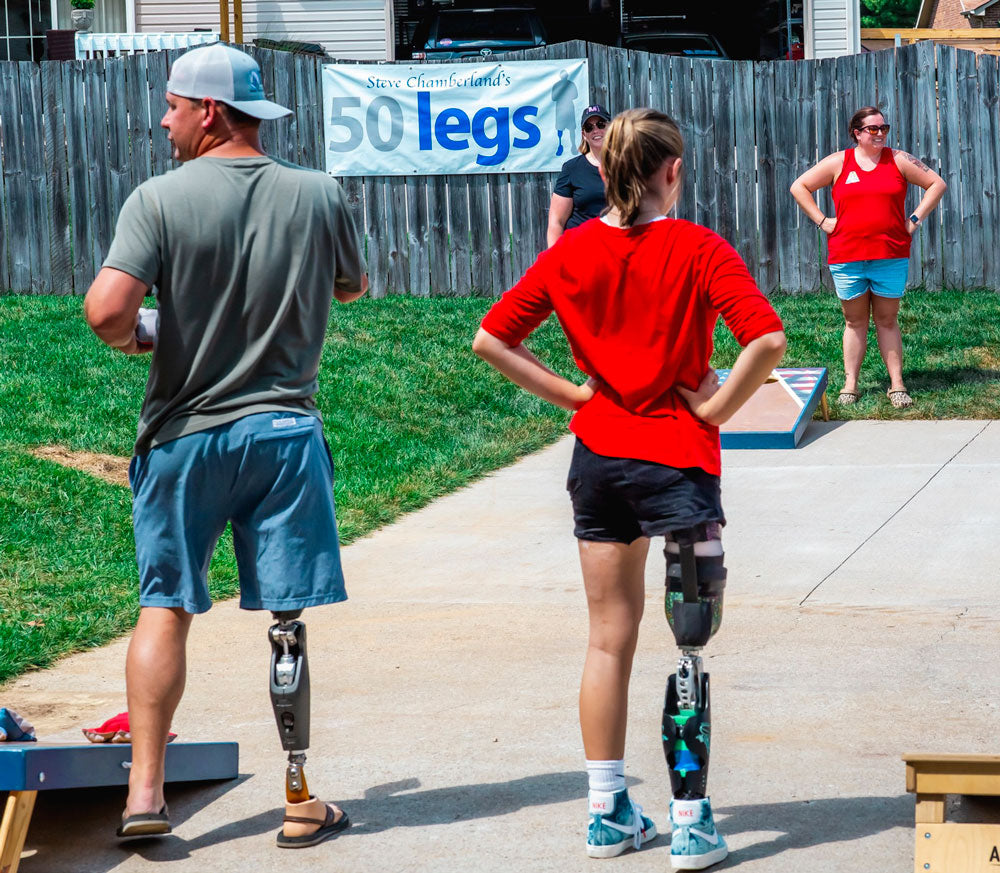 2 RIGHT LEG ABOVE THE KNEE AMPUTEES PLAYING CORNHOLE WITH A 50 LEGS BANNER IN THE BACKGROUND