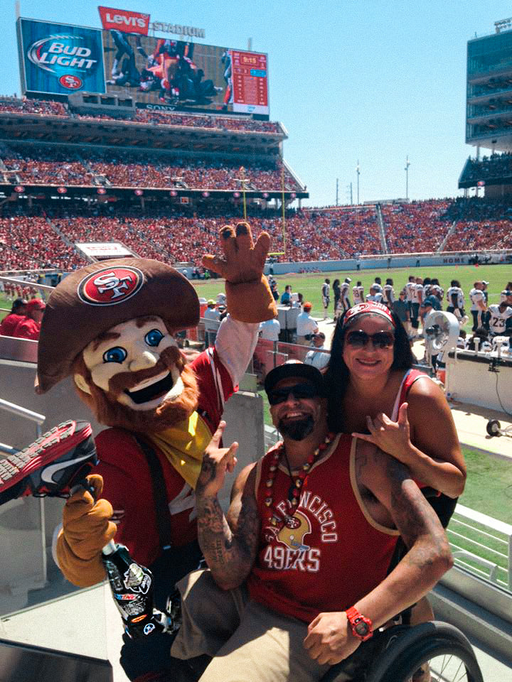 Portrait of Amplife® Foundation & Amplife® Founder Abdul Nevarez with Amplife® Foundation & Amplife® Co-Founder Priscilla Nevarez at the 49ers game with Sourdough Sam holding Abdul's prosthetic leg upward