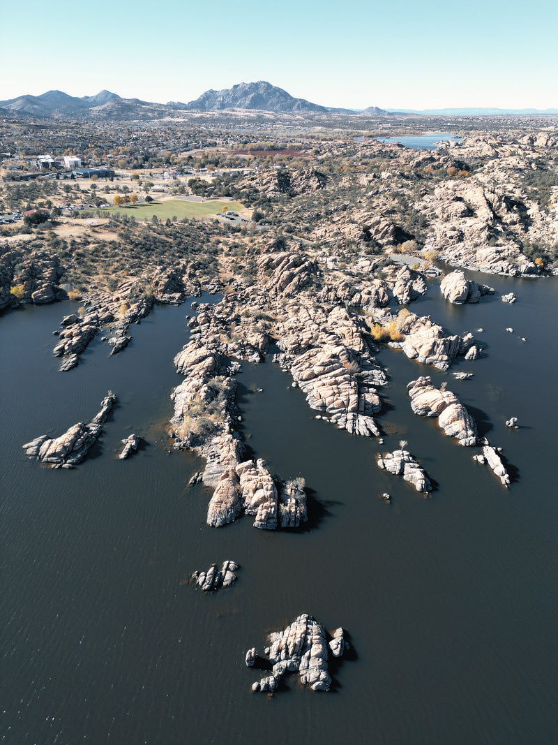 Aerial view of a rocky desert landscape with large stone formations rising from dark water, distant mountains, and a flat valley under a clear blue sky.