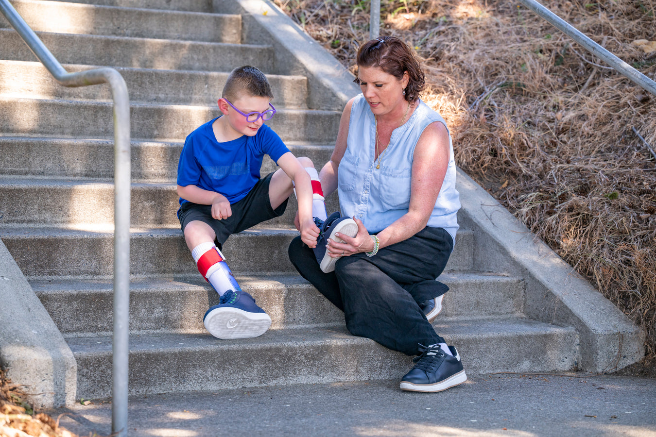 Mother and child sitting on concrete steps outdoors as the child zips up their BILLY Footwear shoes while wearing AFO's with the mother supporting the child's foot on their knee