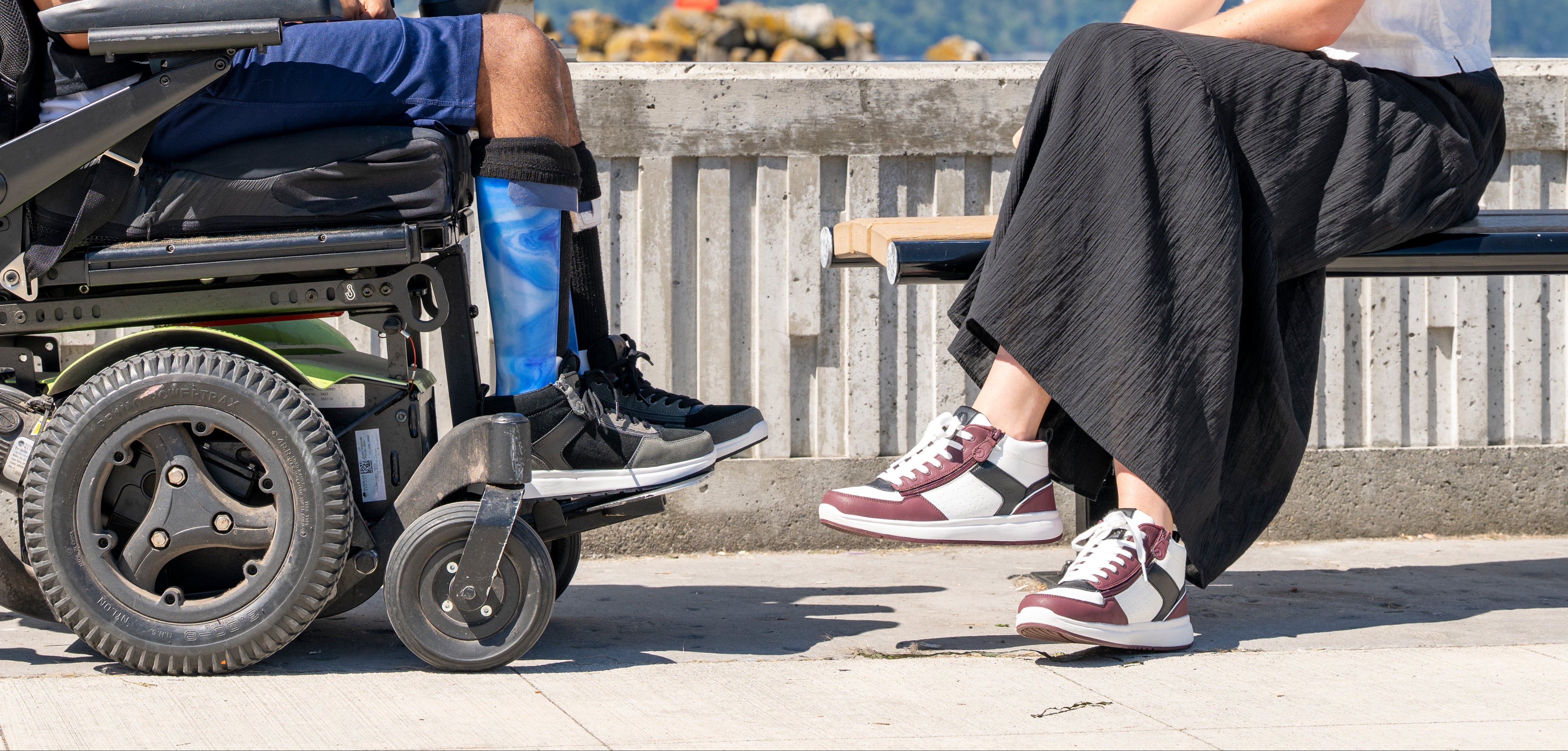 Person using a wheelchair and another person sitting on a bench by a waterfront while both are wearing BILLY Footwear.