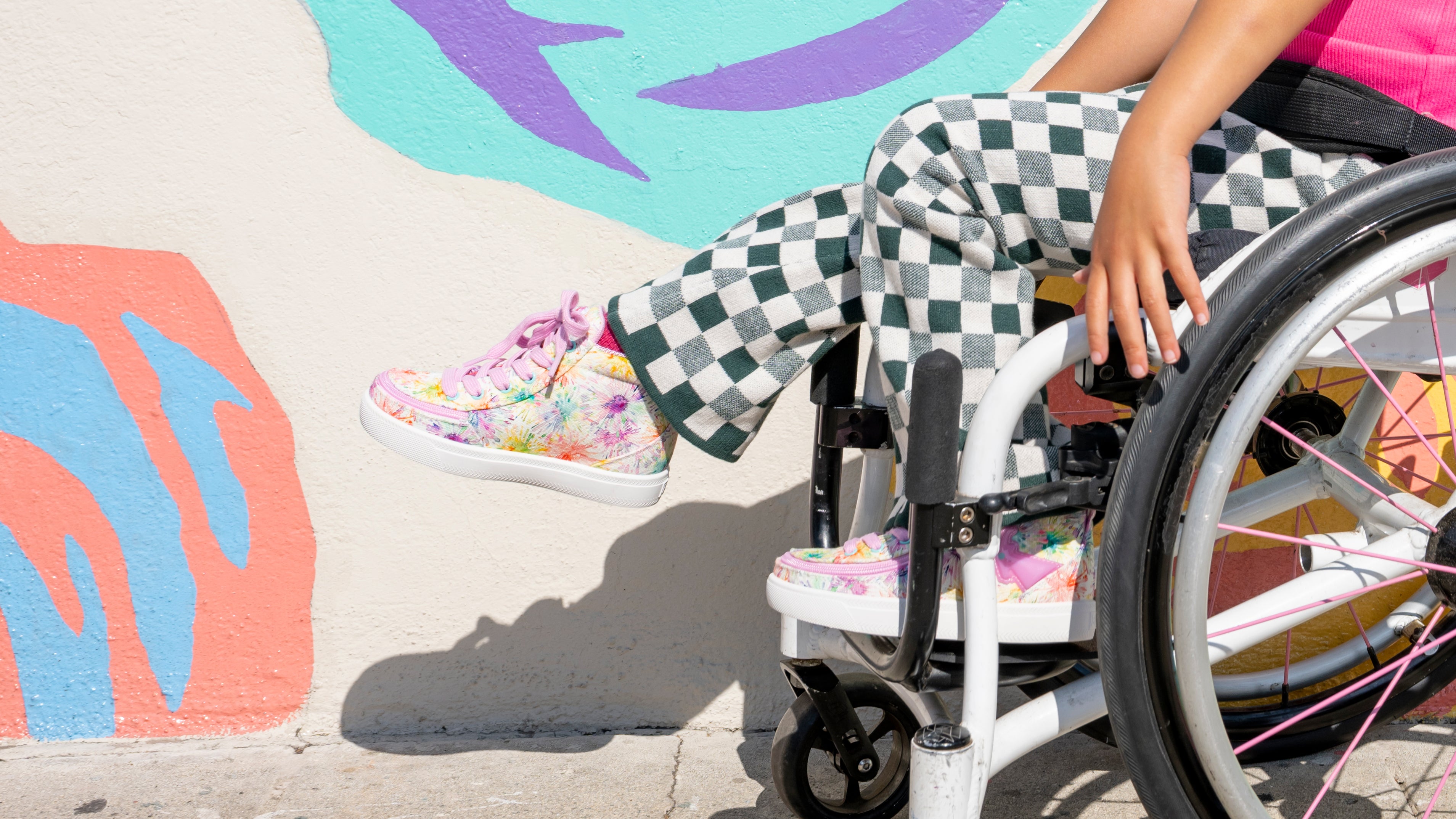 Person in a wheelchair with checkered pants and colorful BILLY Footwear against a colorful wall.