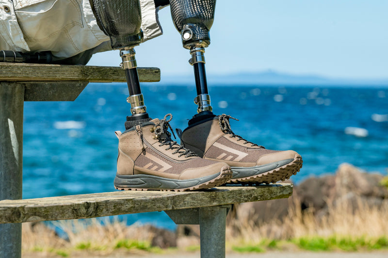 Person with prosthetic legs sitting on a bench by the ocean while wearing BILLY Footwear