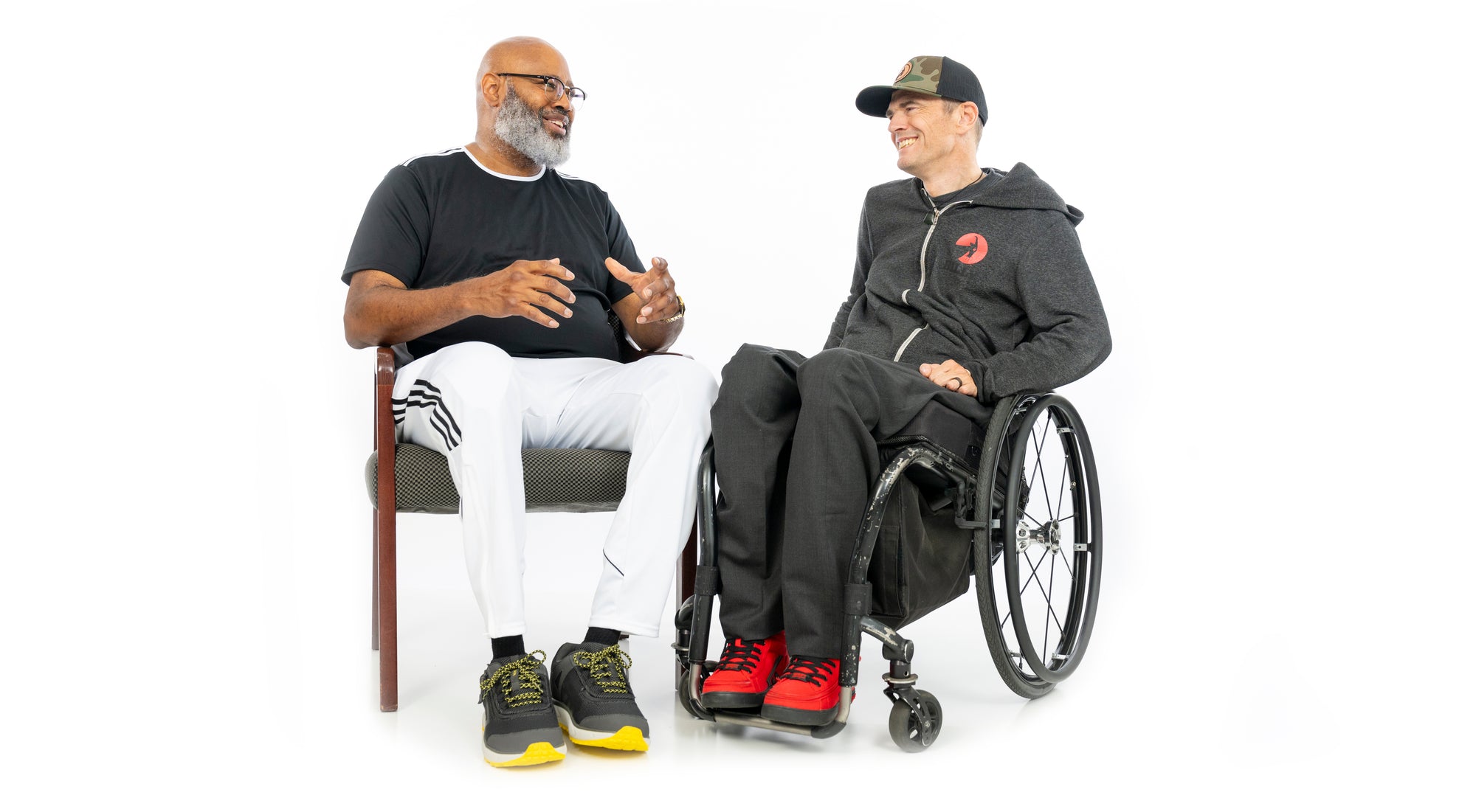 BILLY Footwear Founder Billy Price sitting in his wheelchair discussing with an amputee sitting on a chair while both are wearing BILLY Footwear on a white background