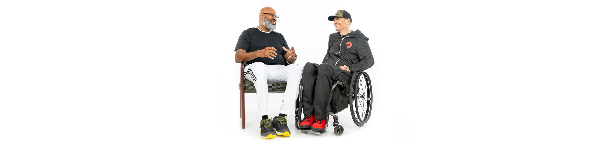 BILLY Footwear Founder Billy Price sitting in his wheelchair discussing with an amputee sitting on a chair while both are wearing BILLY Footwear on a white background