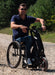 Man riding the Blumil Sport Electric Wheelchair Attachment on a gravel path with greenery in the background