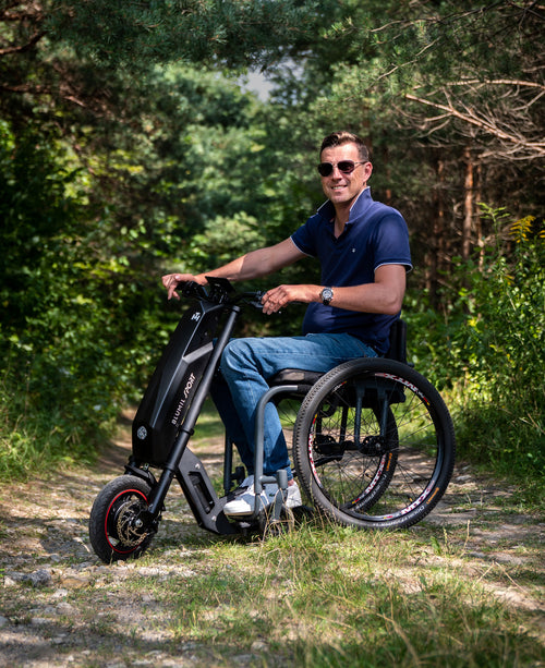 Man riding the Blumil Sport Electric Wheelchair Attachment in a forested area.
