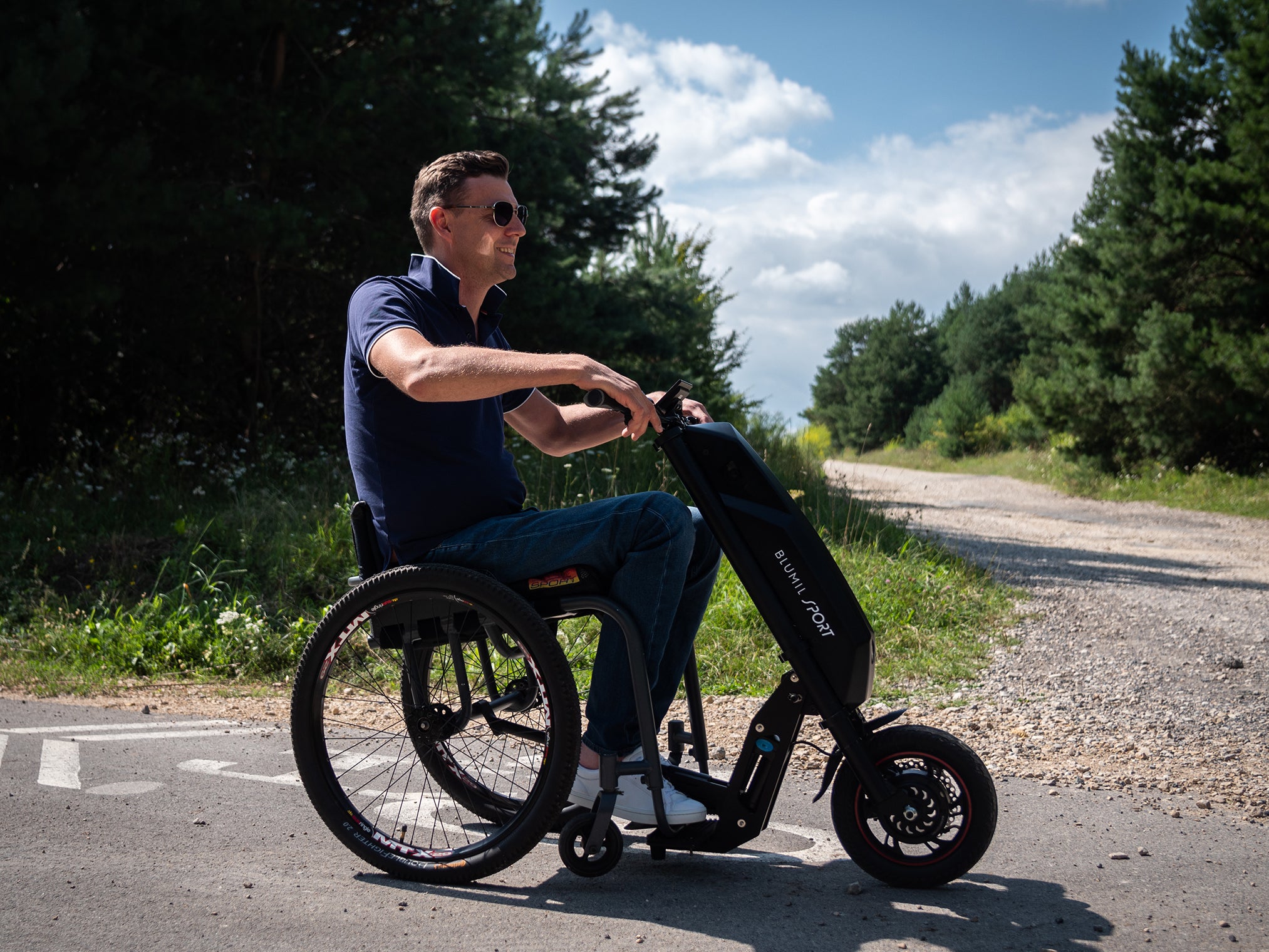 Man riding the Blumil Sport Electric Wheelchair Attachment on a road with trees.
