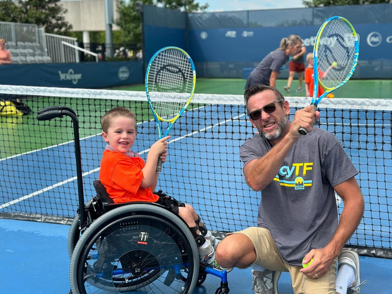 Cincinnati Tennis Foundation Parent and child using a wheelchair posing with tennis racquets on a tennis court.