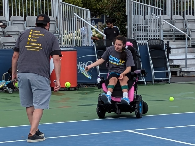Person in a wheelchair playing tennis on a court with another person assisting.