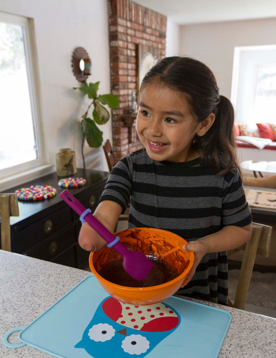 Child holding a bowl of food with a spoon with a blue EaZyHold Grip Aid on a kitchen counter