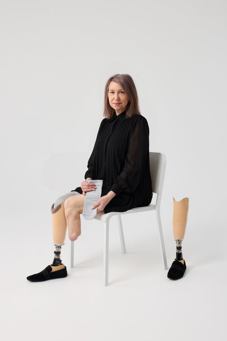 person seated against a white studio background, holding a Ethnocare Underlay Transtibial Closed Prosthetic Sweat Management Device  around their residual limb. A prosthetic lower leg and black shoe are also on the other leg, along with a black shirt on the upper body.
