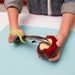 Person using an apple peeler with a green EaZyHold Grip Aid to remove skin from an apple on a light blue surface