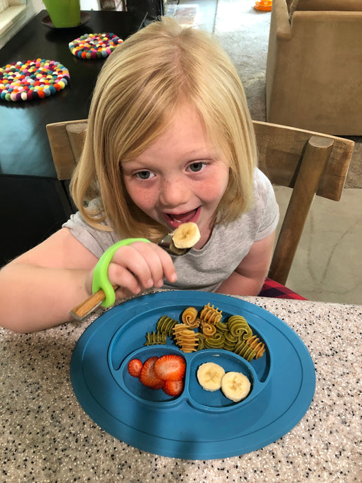 Child eating at a table with a blue plate of food holding the spoon with a green EaZyHold Grip Aid
