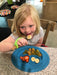 Child eating at a table with a blue plate of food holding the spoon with a green EaZyHold Grip Aid