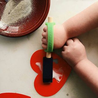 Child's hand holding a black tool with a green EaZyHold Grip Aid over a red heart-shaped object on a light surface