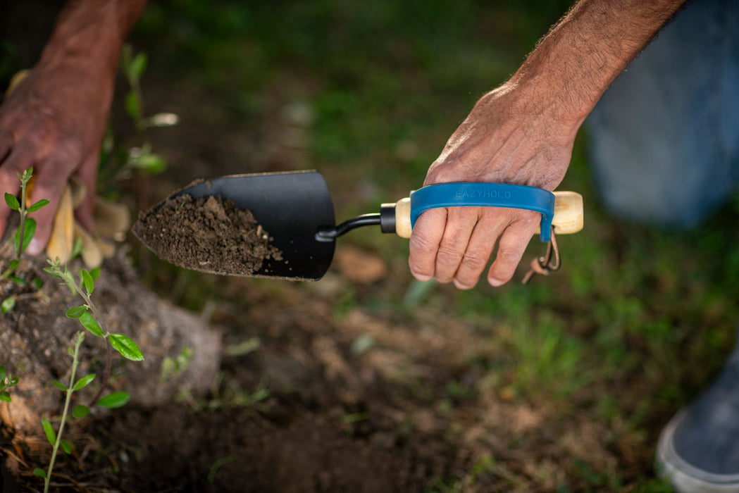 Person holding a trowel with a navy EaZyHold Grip Aid with soil in a garden setting
