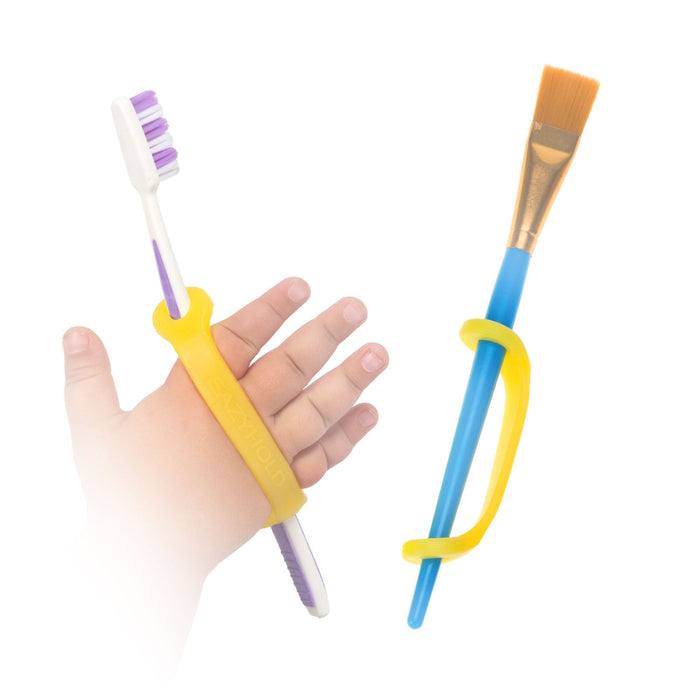Hand holding a toothbrush with a yellow EaZyHold Grip Aid, next to a paintbrush with a yellow EaZyHold Grip Aid on a white background