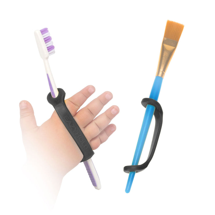 Hand holding a toothbrush with a black EaZyHold Grip Aid, next to a paintbrush with a black EaZyHold Grip Aid  on a white background