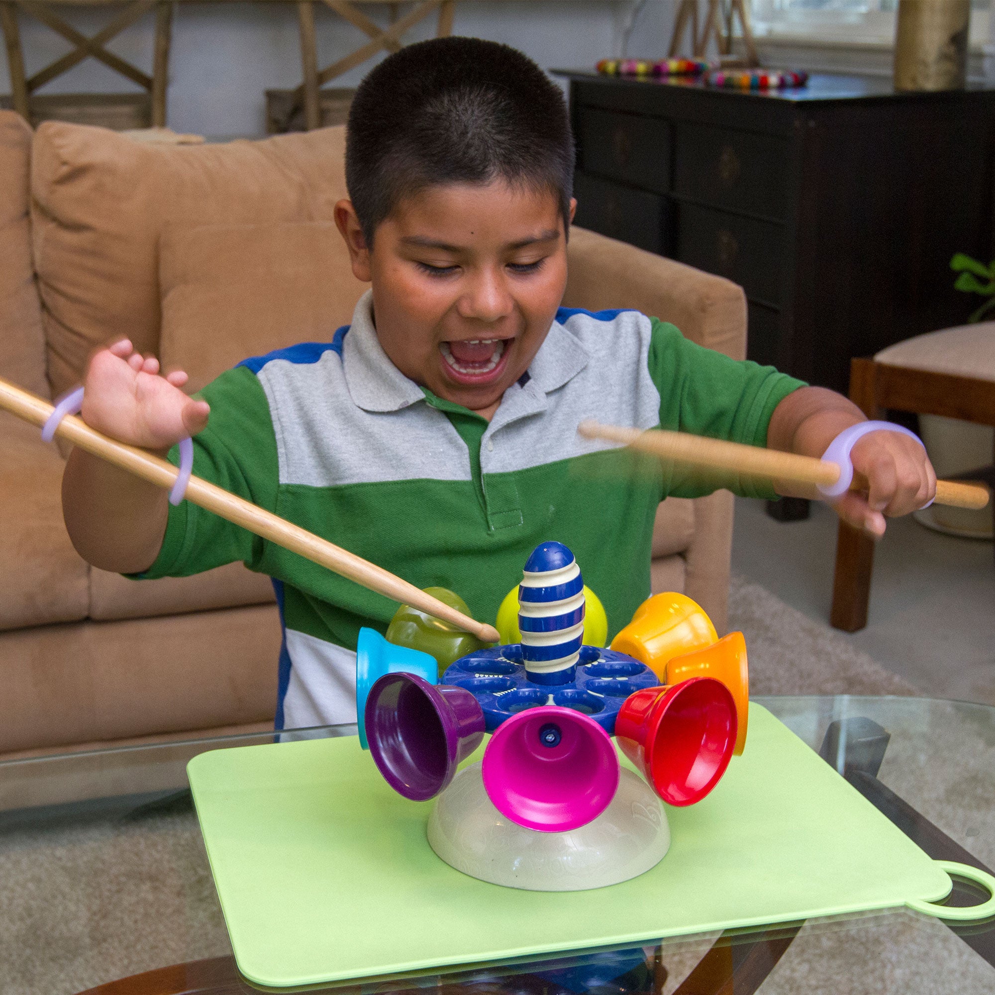Child playing with a colorful instrument on a table in a living room while holding drumsticks with EaZyHold grip aids.