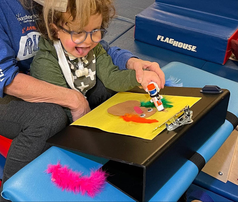 Child and occupational therapist sitting at a table with art supplies, including glue that being held by the functionalhand Universal Cuff Grip Aid