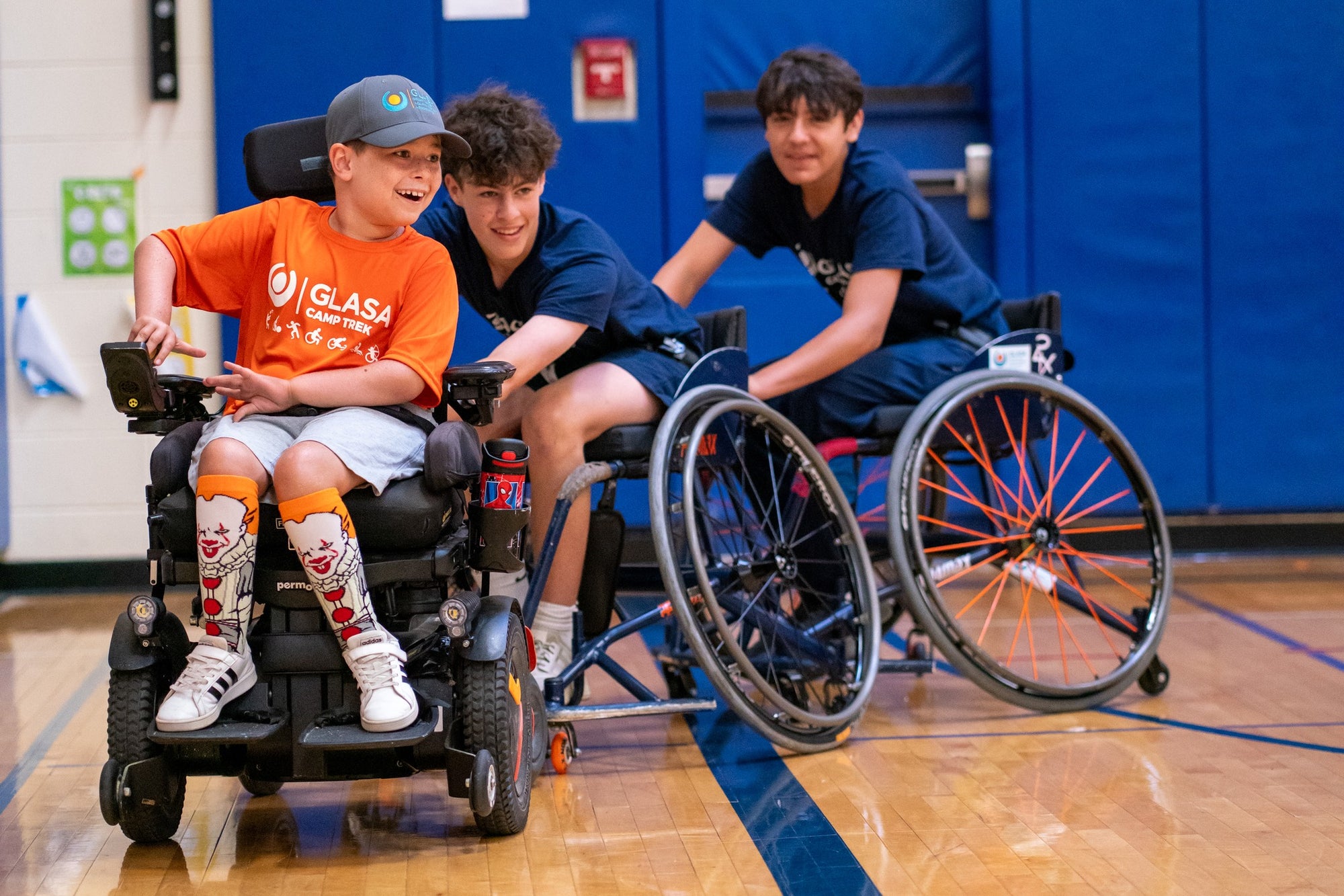 Three young individuals in wheelchairs on a basketball court pushing each other in a train.