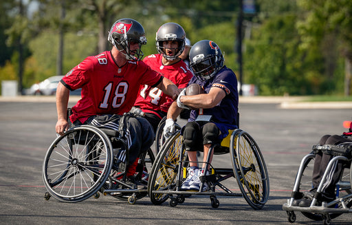 Chicago Bears Wheelchair Football player with the ball rolling past two Tampa Bay Bucs players.