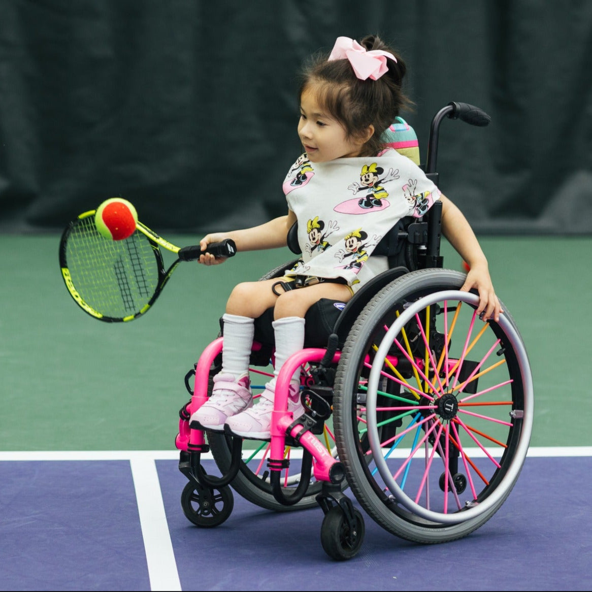 Person in a wheelchair and a child in a pink wheelchair on a tennis court with a tennis racket and ball.