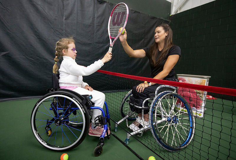 GLASA wheelchair tennis coaching helping a youth player hit the ball on the racquet.