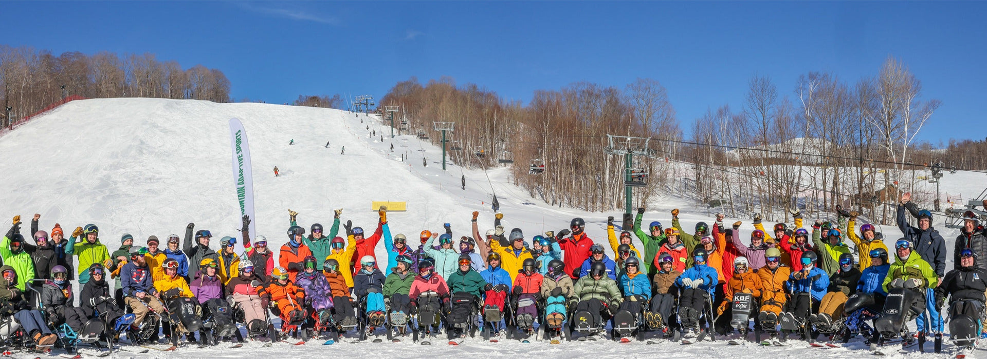 Group of Green Mountain Adaptive Sports participants in colorful winter clothing on a snowy slope with ski lifts and trees in the background