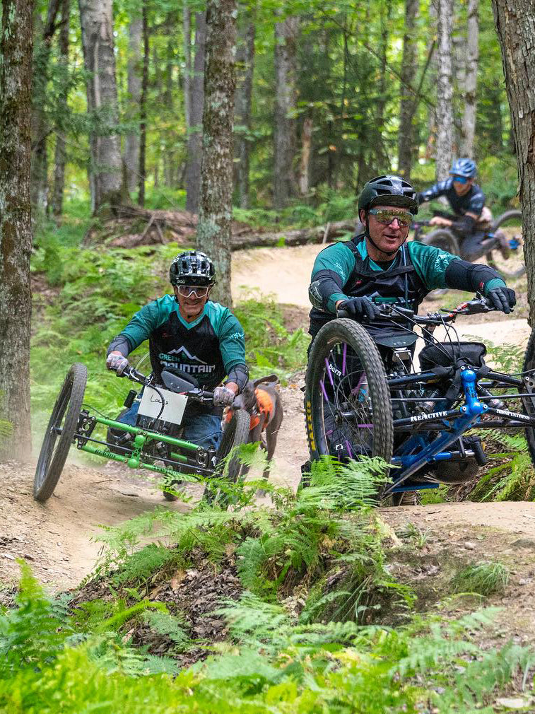 Three individuals on adaptive mountain bikes navigating a forest trail.