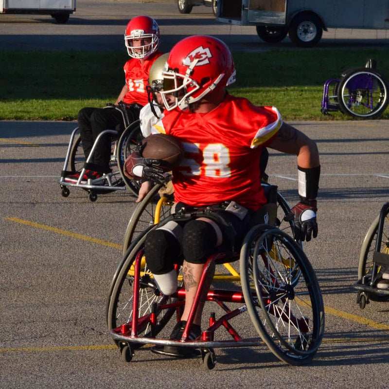 A Kansas City wheelchair football game in progress on an asphalt field, with a player in a red jersey and helmet pushing a wheelchair forward, while other players in wheelchairs compete nearby.