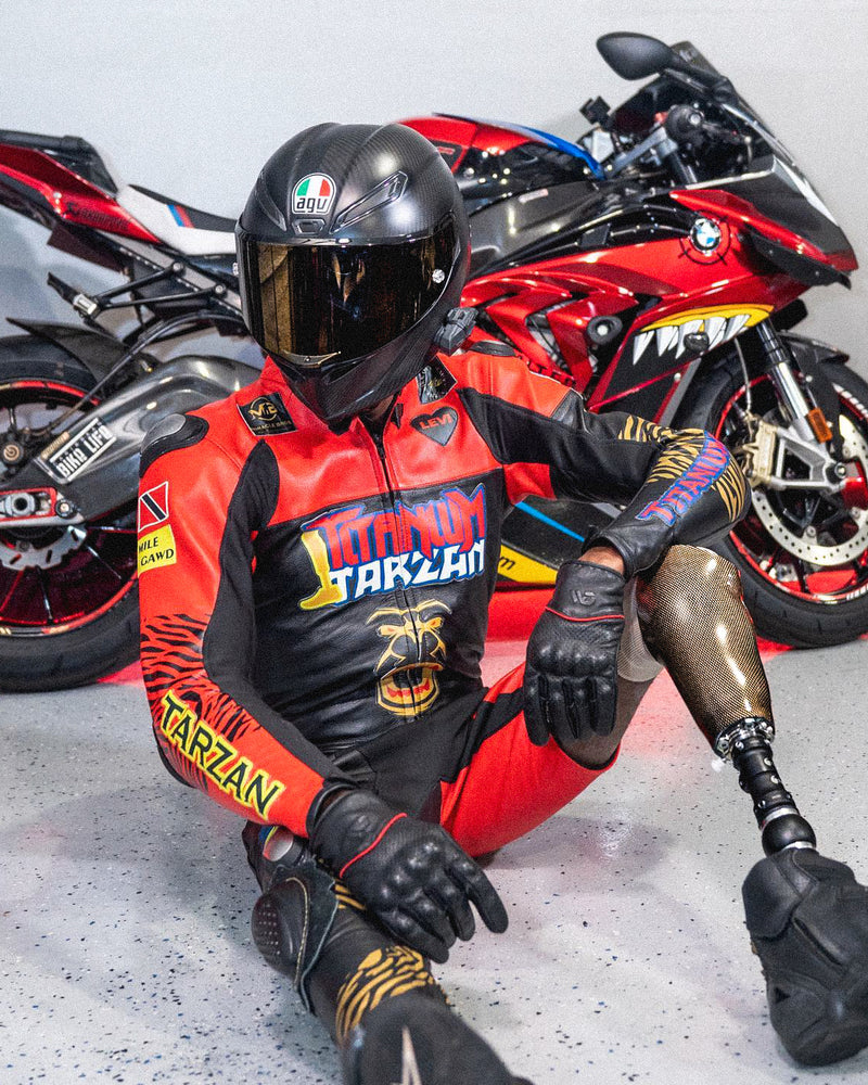 Team Amplife® Ambassador Joseph Forde wearing a black helmet, red and black motorcycle suit with 'Titanium Tarzan' branding, sitting in front of his red motorcycle.