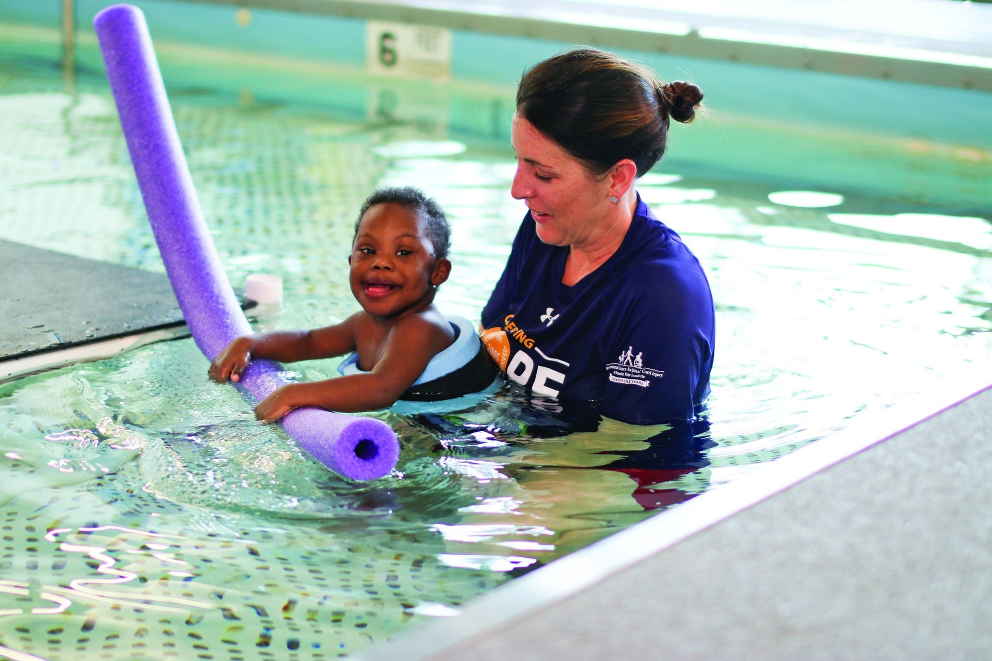 Child and adult from Kennedy Krieger Institute in a pool with a purple pool noodle