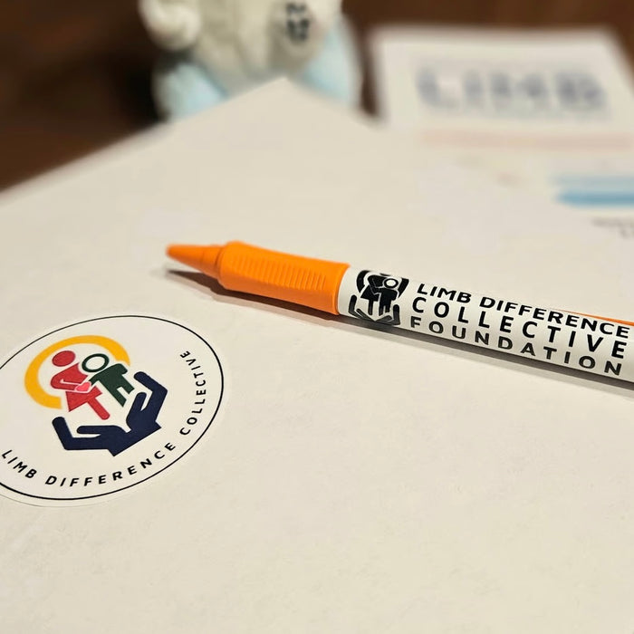 Orange marker with 'Limb Difference Collective Foundation' branding laid on a piece of paper with a sticker featuring a similar logo