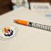 Orange marker with 'Limb Difference Collective Foundation' branding laid on a piece of paper with a sticker featuring a similar logo