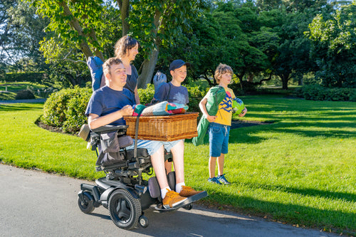 Person using a power wheelchair holding a basket with LapStacker, surrounded by family in a park setting.