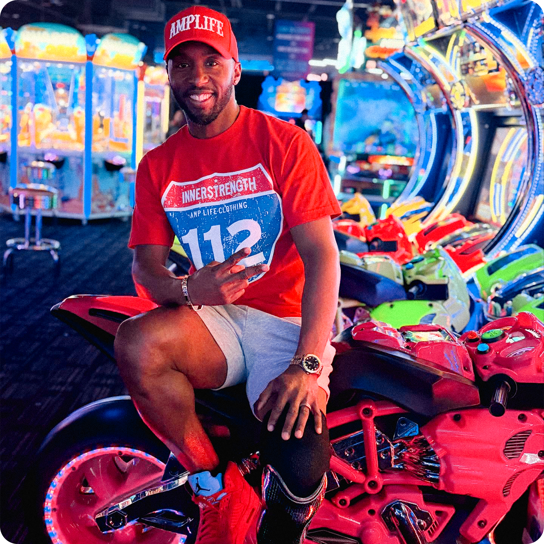 Team Amplife® Ambassador Michael Braxton Jr. wearing the Amplife® Curved Bill Snapback & Amplife® Innerstrength 112 Sign T-Shirt while sitting on an arcade motorcycle