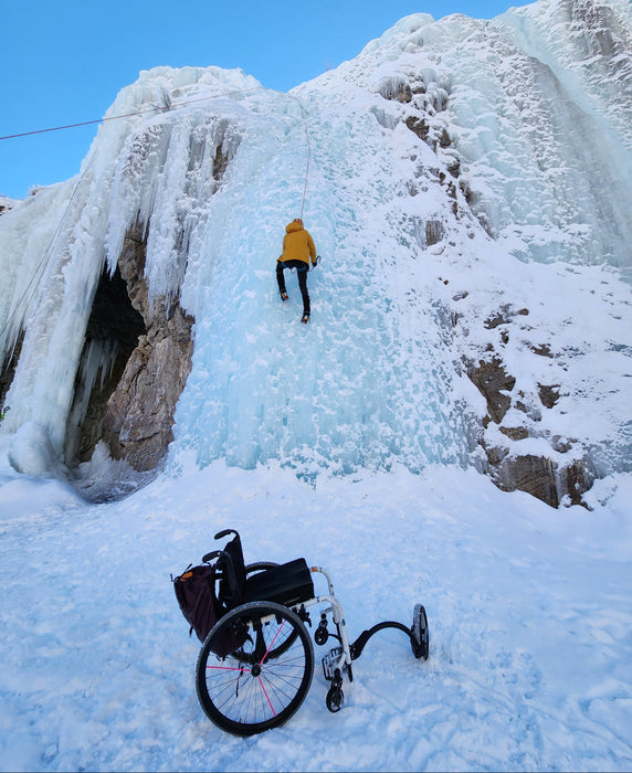 Person ice climbing a frozen waterfall with a wheelchair left on the snow below