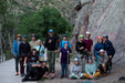 Group photo of Paradox Sports climbers from an outdoor meet up at Clear Creek Canyon. The group is smiling at the camera and the rock wall is to the right and the creek path and creek are to the left.