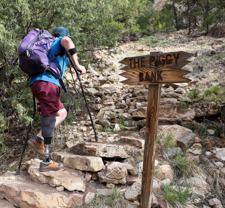 Hiker with a prosthetic leg on a rocky trail next to a signpost with 'The Piggy Bank' written on it.